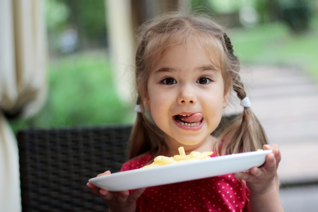Beautiful laughing little girl sitting at table and holding a plate with French fries, food and drink concept, outdoor portrait Beautiful laughing little girl sitting at table and holding a plate with French fries, food and drink concept, outdoor portrait