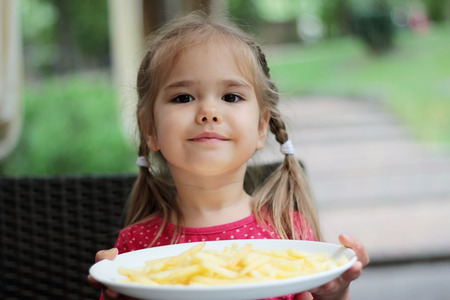 Beautiful laughing little girl sitting at table and holding a plate with French fries, food and drink concept, outdoor portraitの写真素材