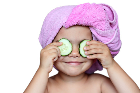 Funny small girl with cucumbers on eyes and a bath towel on her head over white background, beauty and health concept, indoor closeup portraitの写真素材