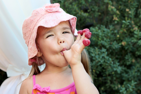 Cute little girl holding raspberries on her fingers, summertime outdoor, healthy food conceptの写真素材