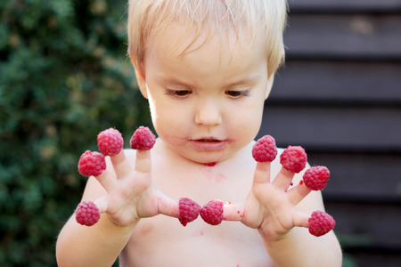 Cute dirty toddler boy holding raspberries on his fingers, summertime outdoor, healthy food conceptの写真素材