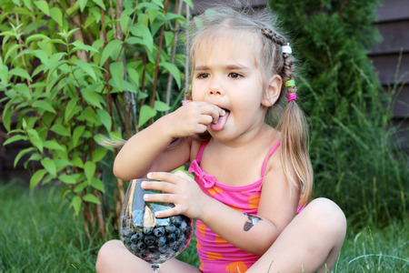 Cute little girl sitting on the grass and eating blueberries from the glass, summertime outdoor, healthy food conceptの写真素材