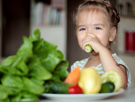 Happy little girl eating cucumber from a plate full of fresh vegetables, healthy food concept, indoor portraitの写真素材
