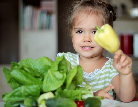 Cute little girl playing with pepper before a plate full of fresh vegetables, healthy food concept, indoor portraitの写真素材