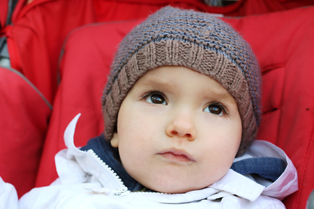 Thoughtful baby in the knitted hat sitting in the stroller, outdoor portraitの写真素材