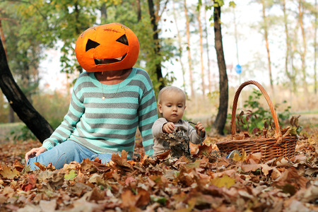 Handsome toddler sitting on the leaves near the adult in pumpkin mask on her head in the autumn parkの写真素材