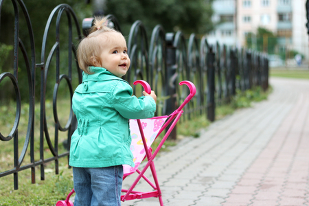 Stylish little child wearing green coat, pushing toy stroller and looking around, outdoors in the city parkの写真素材