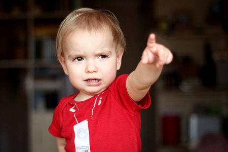 Serious toddler boy wearing in a red T-shirt points a finger to something, people gesture concept, emotional indoor portraitの写真素材