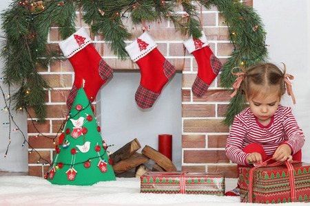 Pretty child girl opening a gift near Christmas decorated fireplace, winter holiday family conceptの写真素材