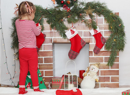 Pretty child girl looking for a gift in Santa sock near Christmas decorated fireplace, winter holiday family conceptの写真素材