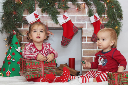 Two cute toddlers (boy and girl) sitting near Christmas decorated fireplace, winter holiday family conceptの写真素材