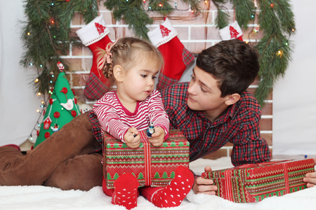 Two children (teenager boy and toddler girl) looking at each other near gifts and Christmas decorated fireplace, winter holiday family conceptの写真素材
