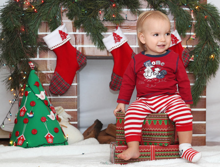 Smiling cute toddler boy sitting on a packed gift and looking for Santa near Christmas decorated fireplace, winter holiday family conceptの写真素材