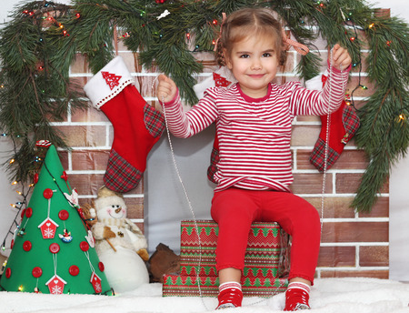 Pretty child girl sitting on packed gifts and holding a garland decoration near Christmas decorated fireplace, winter holiday family conceptの写真素材