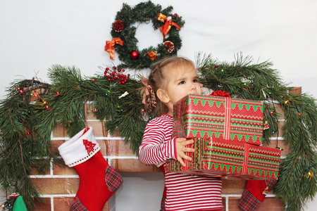 Pretty child girl holding some packed gifts near Christmas decorated fireplace, winter holiday family conceptの写真素材