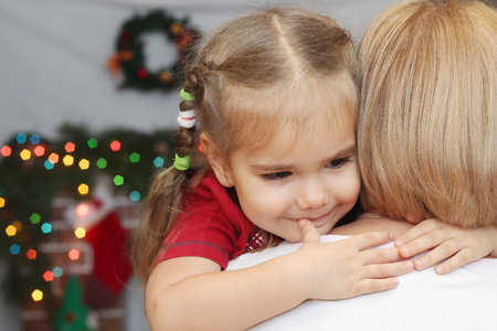 Cute little girl hugging the adult woman (back view) over blurred Christmas decorated fireplace on background, winter holiday family conceptの写真素材