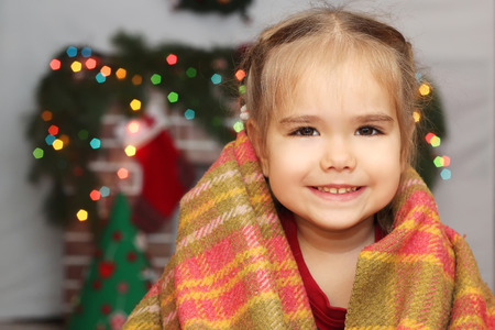Happy little girl covering herself up in blanket over blurred Christmas decorated fireplace on background, winter holiday family conceptの写真素材