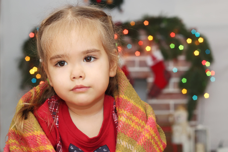 Cute little girl covering herself up in blanket over blurred Christmas decorated fireplace on background, winter holiday family conceptの写真素材