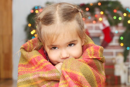 Happy little girl covering herself up in blanket over blurred Christmas decorated fireplace on background, winter holiday family conceptの写真素材