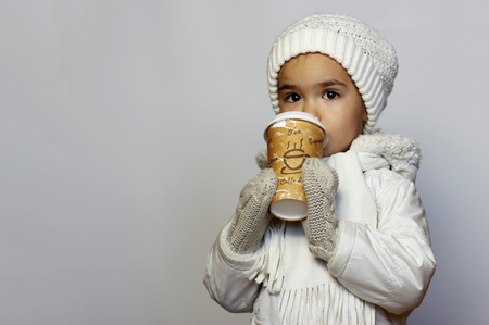Cute little girl holding mug and drinking tea in white coat, white knitted hats, scarves and mittens over white background, winter outfitの写真素材
