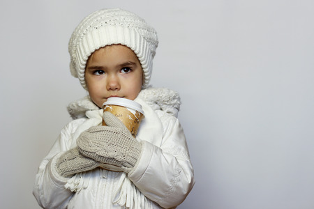 Cute little girl holding mug and drinking tea in white coat, white knitted hats, scarves and mittens over white background, winter outfitの写真素材