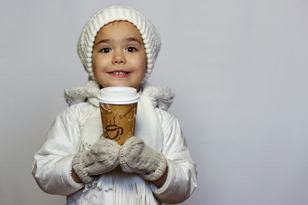 Cute little girl holding mug and drinking tea in white coat, white knitted hats, scarves and mittens over white background, winter outfitの写真素材