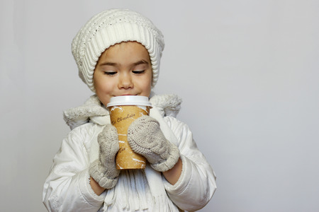 Cute little girl holding mug and drinking tea in white coat, white knitted hats, scarves and mittens over white background, winter outfitの写真素材