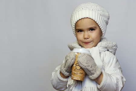 Cute little girl holding mug and drinking tea in white coat, white knitted hats, scarves and mittens over white background, winter outfitの写真素材