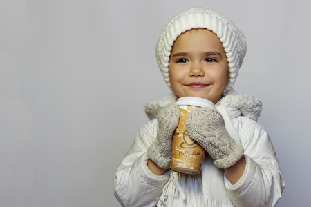 Cute little girl holding mug and drinking tea in white coat, white knitted hats, scarves and mittens over white background, winter outfitの写真素材