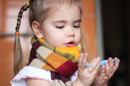 Young sad child (girl) with a thermometer in her hands measuring the fever, indoor portraitの写真素材