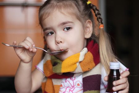 Young sad child (girl) in the scarf keeping a spoon with liquid syrup and mixture for flu, indoor portraitの写真素材