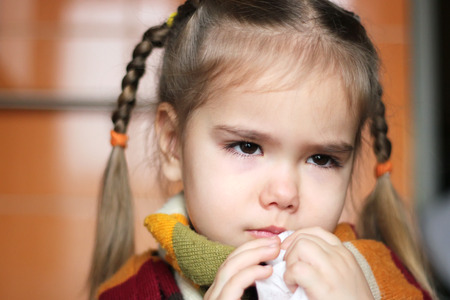 Sad young child (girl) with sick look and in the scarf on her throat blowing her nose in the napkin, indoor portraitの写真素材