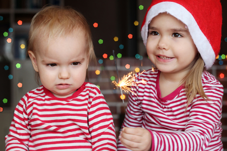Handsome toddler boy and cute small girl in Santa hat hold burning sparkler and smile happily over defocused Christmas backgroundの写真素材