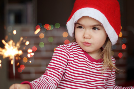 Happy small girl in Santa hat squints slyly at burning sparkler in her hand over Christmas backgroundの写真素材