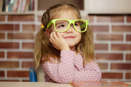 Portrait of a pretty child girl in funny green eyeglasses sitting at the desk with a book, indoor education conceptの写真素材