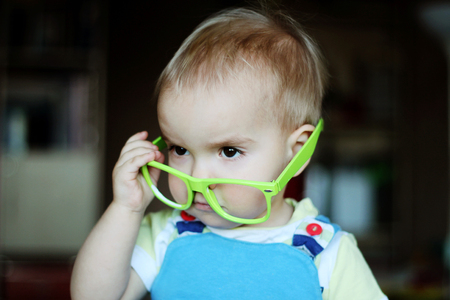 Portrait of cute kid boy with green eyeglasses holding glasses with hand and looking attentively, indoor portrait, educational conceptの写真素材