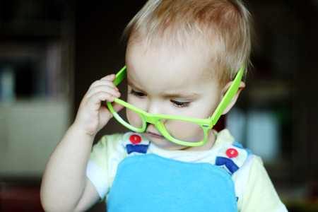 Portrait of cute kid boy with green eyeglasses holding glasses with hand and looking attentively, indoor portrait, educational conceptの写真素材