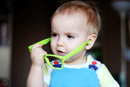 Portrait of cute kid boy with green eyeglasses holding glasses with hand and looking attentively, indoor portrait, educational conceptの写真素材
