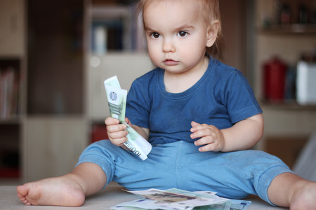 Toddler boy squeezes a denomination (paper note) sitting among wad of money, indoor financial conceptの写真素材