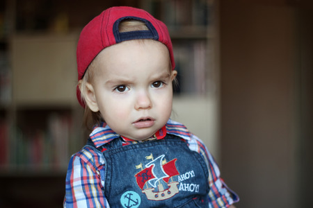 Handsome toddler boy looks seriously wearing in jeans overalls and a red cap, indoor portrait, building conceptの写真素材