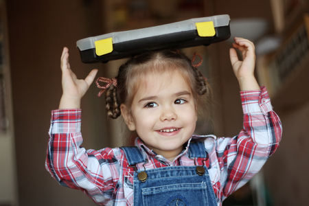 Pretty child girl wearing in jeans overalls holds a set of tools on her head, indoor portrait, building conceptの写真素材