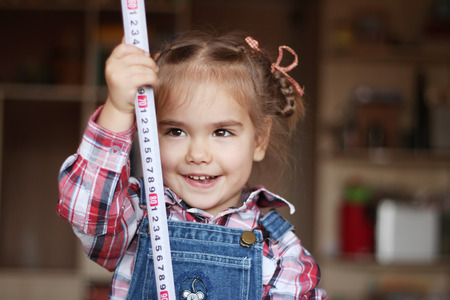 Pretty child girl wearing in jeans overalls holds a measuring tape in her hands, indoor portrait, building conceptの写真素材