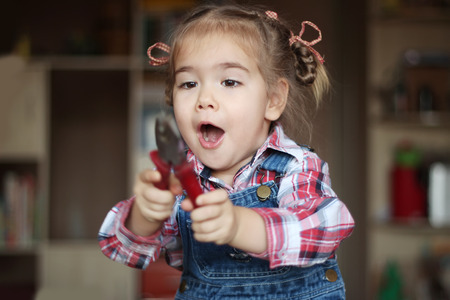 Pretty child girl wearing in jeans overalls holds flat nose pliers and works with inspiration, indoor portrait, building conceptの写真素材