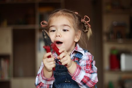 Pretty child girl wearing in jeans overalls holds flat nose pliers and works with inspiration, indoor portrait, building conceptの写真素材