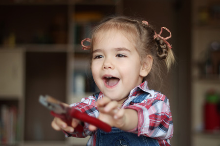 Pretty child girl wearing in jeans overalls holds flat nose pliers and works with inspiration, indoor portrait, building conceptの写真素材