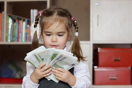 Inspired little child girl wearing in grey business style dress holds hundred Euro denominations (paper money) like a fan, indoor financial conceptの写真素材