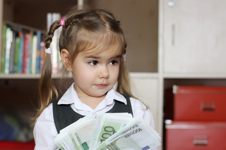 Surprised little child girl wearing in grey business style dress holds hundred Euro denominations (paper money) like a fan, indoor financial conceptの写真素材