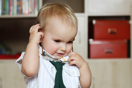 Handsome toddler boy wearing in a white shirt with a green tie takes away his eyeglasses, business concept, indoor portraitの写真素材