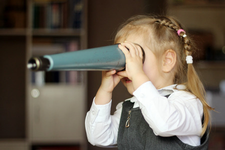 Pretty child girl wearing in grey dress and white shirt looks forward through spyglass, education concept, indoorの写真素材