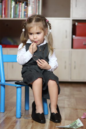Little child girl wearing in grey business style dress takes off big black shoes with heels and bites an ear of eye-glassesの写真素材